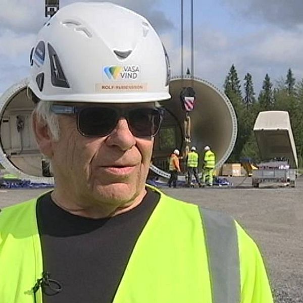 Bild på Rolf Rubensson  klädd i varselkläder och bygghjälm framför arbetare som reser ett vindkraftverk i Åskälen