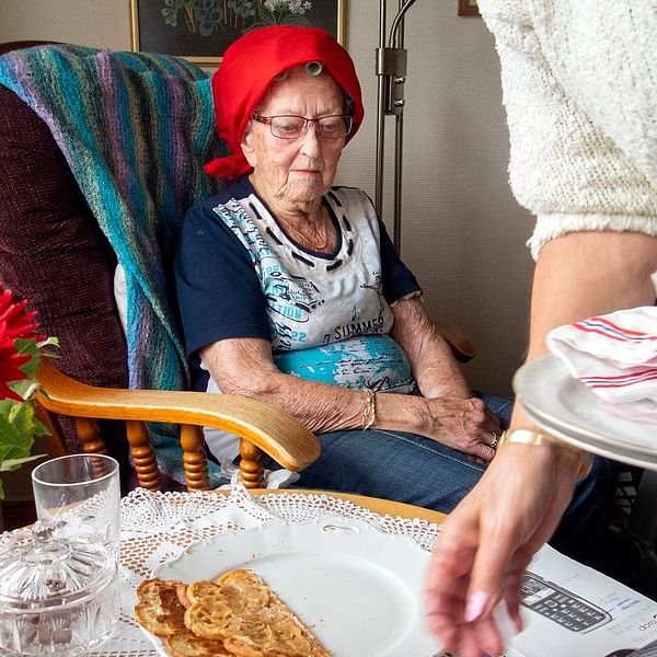 Äldre dam sitter vid ett bord och hemtjänstpersonal plockar bort disk