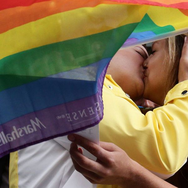 Två personer kysser varandra bakom en regnbågsflagga.