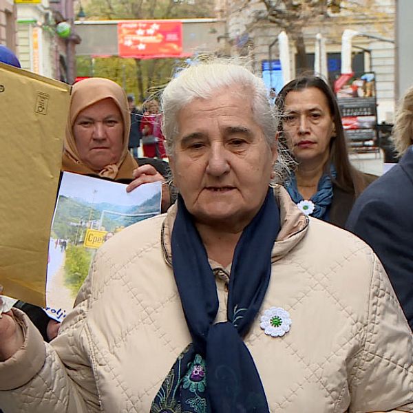 Flera personer står på en gata och protesterar. I mitten en äldre dam som håller upp ett stort kuvert.