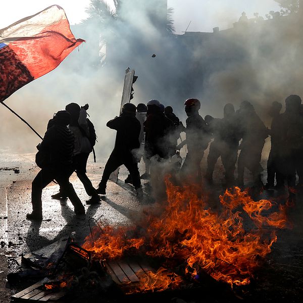 Demonstranter i Chile håller upp en chilensk flaga och står intill bråte som står i lågor.