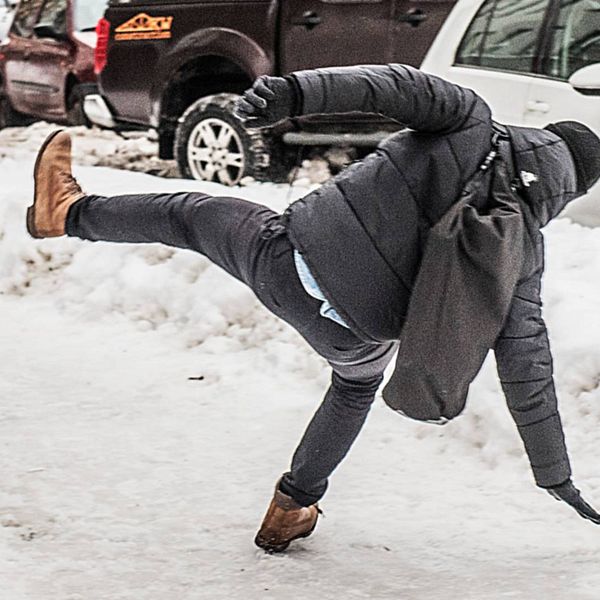 En man iklädd svarta vinterkläder och ryggsäck halkar på en snöbeklädd gata.