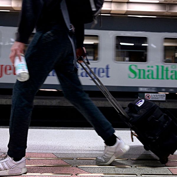 Person går med rullväska på perrong. I bakgrunden ett Snälltåg.