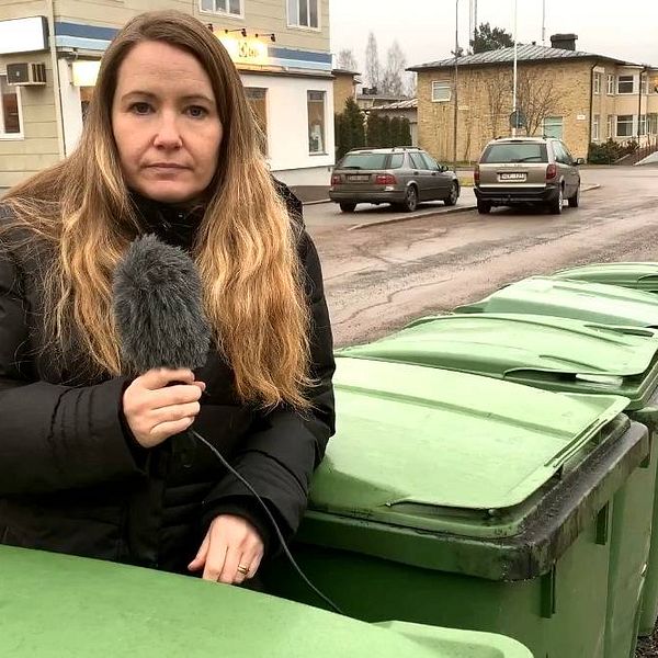 kvinna håller i mick och står framför flera gröna soptunnor.