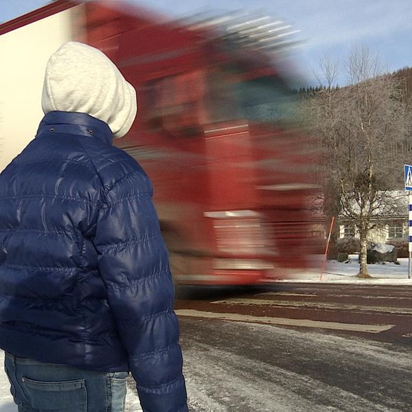 Barn i blå jacka står vid ett övergångsställe där en lastbil precis kör förbi.