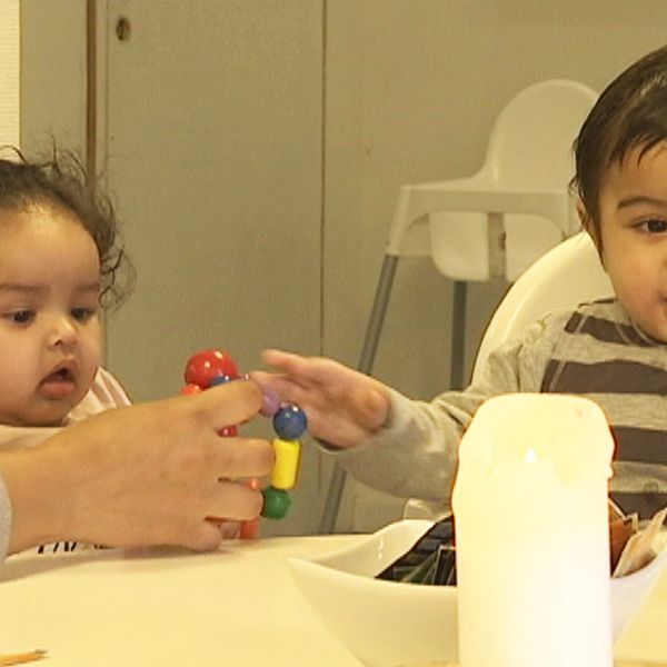 Två småbarn sitter vid ett bord och leker med leksaker. En vuxen hand ger en leksak till ett av barnen.
