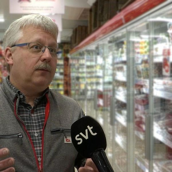 Man med mustasch och glasögon står i en matbutik med en mikrofon framför ansiktet.