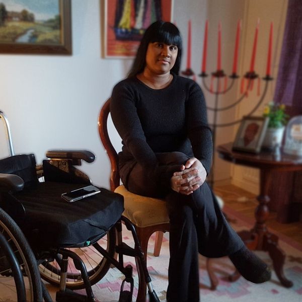 Kvinna sitter på en stol i ett vardagsrum med en rullstol bredvid sig.