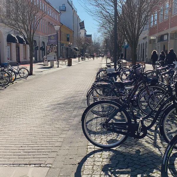 En folktom gågata med cyklar, parkerade i cykelställ, i förgrunden.