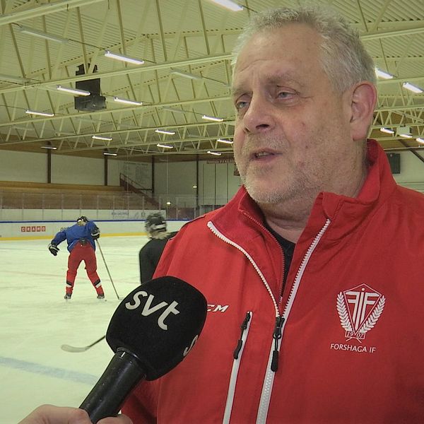 Christer Larsson i röd jacka blir intervjuad vid sidan av hockeyrinken. Tre spelare syns i bakgrunden.