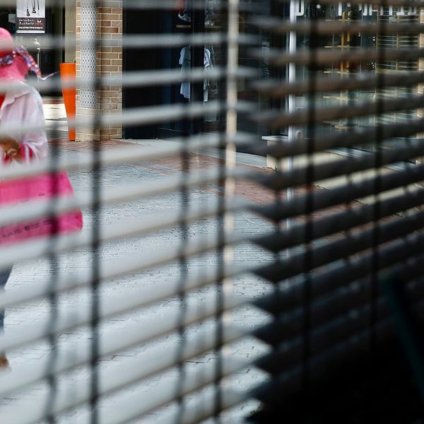En kvinna kollar i sin mobil och iakttas genom ett fönster med halvdragna persienner.