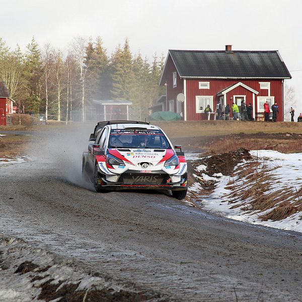 En rallybil på en grusväg med lite snö vid sidan. I bakgrunden syns lador och en röd stuga med vita knutar.