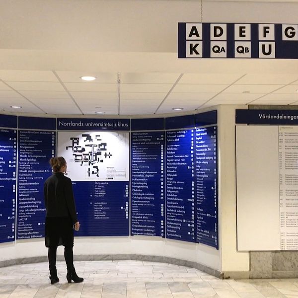 En kvinna i mörka kläder står och tittar på orienteringstavland i centralhallen på Norrlandsuniversitetsjukhus. I förgrunden syns skyltar och pilar som är upphängda från taket.