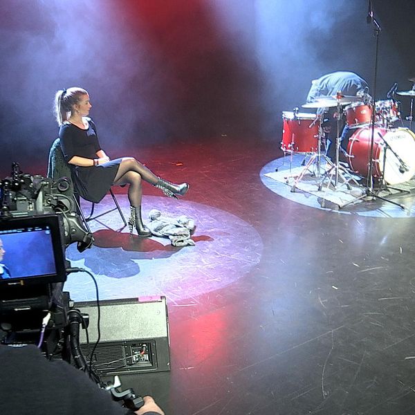 Emmy sitter på en stol på scenen, bredvid ett trumset. I förgrunden en tv-kamera som filmar henne.