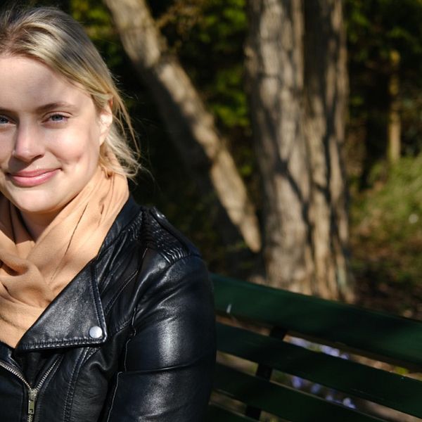 Hanna sitter på en parkbänk, i bakgrunden syns lummig grönska.