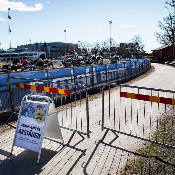 I förgrunden en skylt som visar att publikplatserna är avstängda, och i bakgrunden kör ekipagen i ett travlopp förbi på Färjestadstravet i Karlstad.