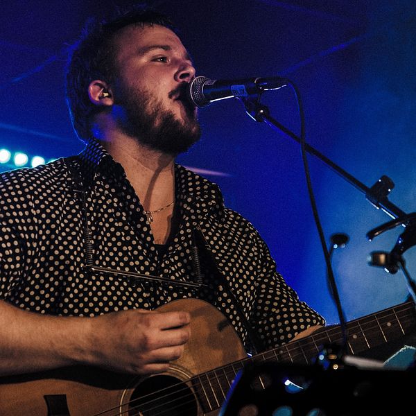 Trubaduren Christopher Edgar står på scenen i blått ljus och spelar gitarr och sjunger iklädd en svart skjorta med vita prickar.