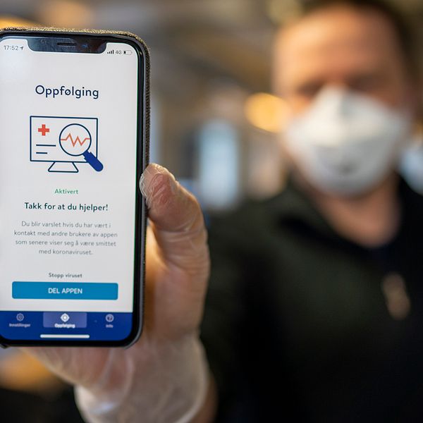 En man med ansiktsmask syns i bakgrunden. Han har plasthandskar på sig och håller upp en mobiltelefon där appen ”Smittestopp” syns.