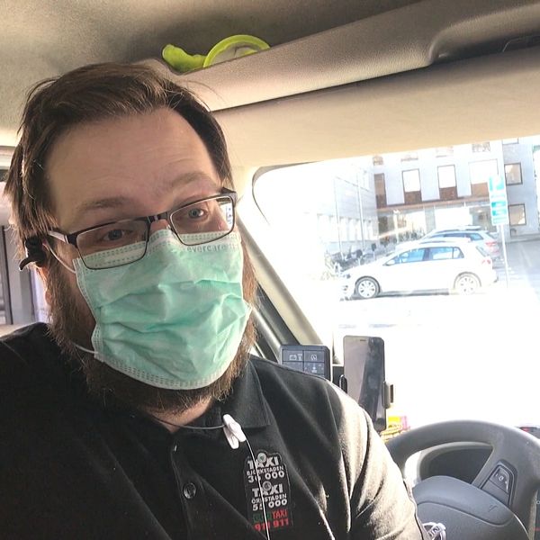 En taxichaufför med glasögon och munskydd som sitter vid ratten i sin bil utanför Norrlands universitetssjukhus i Umeå. Han har svart pikétröja.