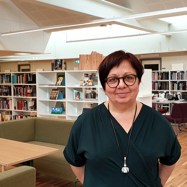 En kvinna med kort mörkbrunt hår, glasögon och grön tröja tittar in i kameran. Hon står i ett skolbibliotek.