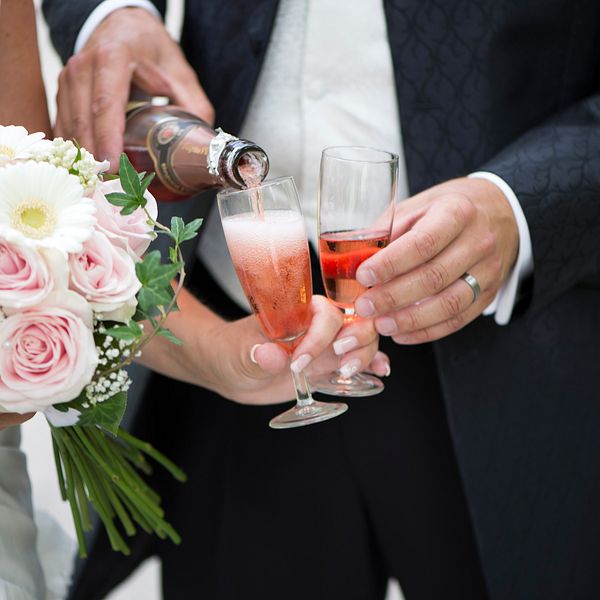 Närbild på brudpar, beskuren från axlarna och ned. Från vänster: bruden håller i en bukett av rosor och prästkragar medan brudgummen häller upp rosa champagne i hennes champagneglas.