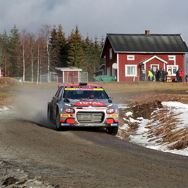 En rallybil på en grusig väg med en röd stuga samt lador i bakgrunden.