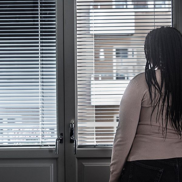 Genrebild. En kvinna står och tittar ut genom ett fönster med persienner.
