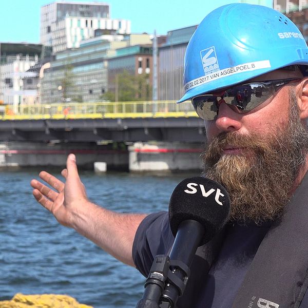 Man med blå bygghjälm står invid vattnet med centrala Stockholm som fond. Han pekar mot den bro som monteras bakom honom.