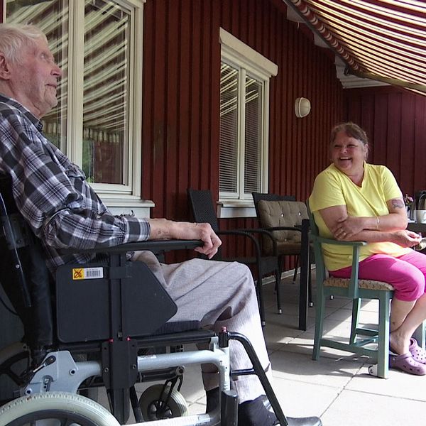 En äldre man i rullstol och en kvinna i färgglada kläder sitter och samtalar