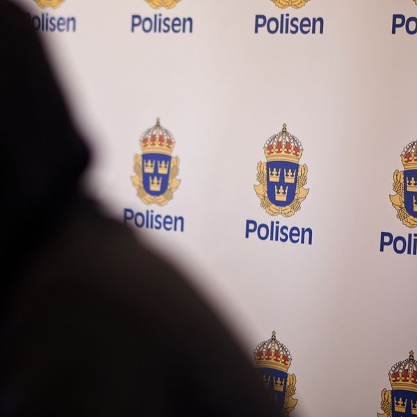 En oskarp bild som visar en del av en person i profil framför en bakgrund med polisens logotyp och texten ”Polisen”.