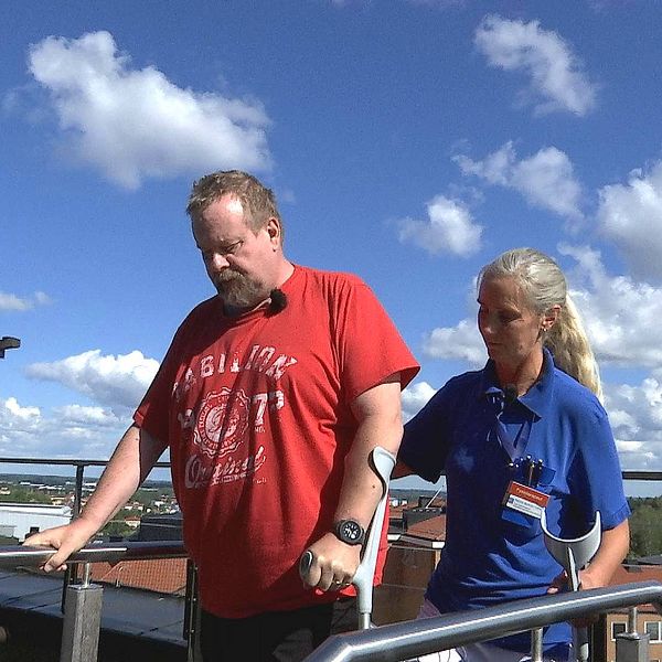 Johan står på en liten trappa uppe på en takterass med en molning himmel bakom. Han har kryckor och ansiktet är sammanbitet. Bredvid honom står fysioterapeuten Marina med en hand på hans rygg.