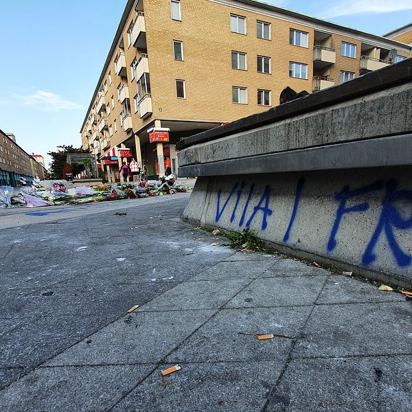 Någon har klottrat ”Vila i frid” på en sittplats vid Nyforsgatan. I bakgrunden syns flerfamiljshus i gult tegel och ett hav av blommor och ljus.