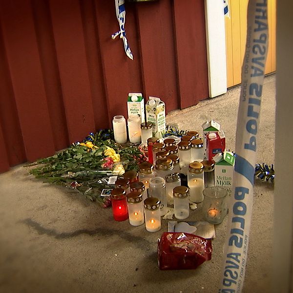 Ljus, blommor och mjölkförpackningar står utanför en lägenhetsdörr, avspärrningsband från polisen syns