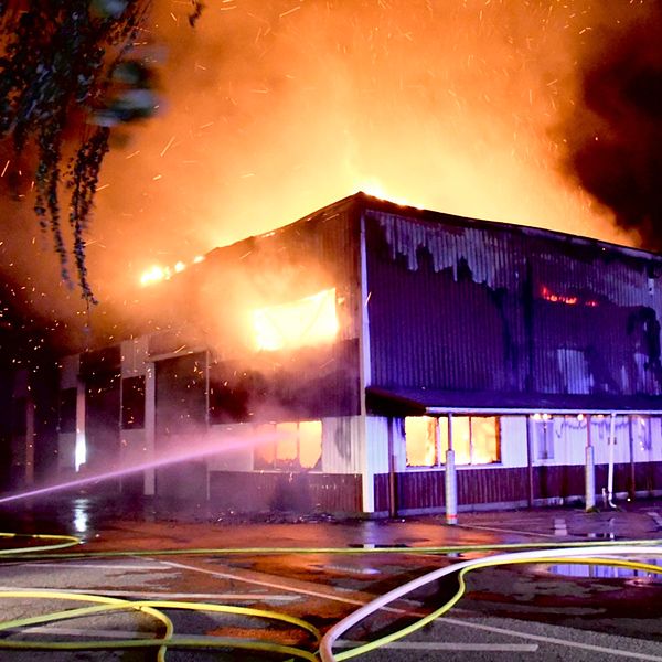 Ett fyrkantigt hus som brinner och en person står på knä med en brandslang som sprutar vatten.
