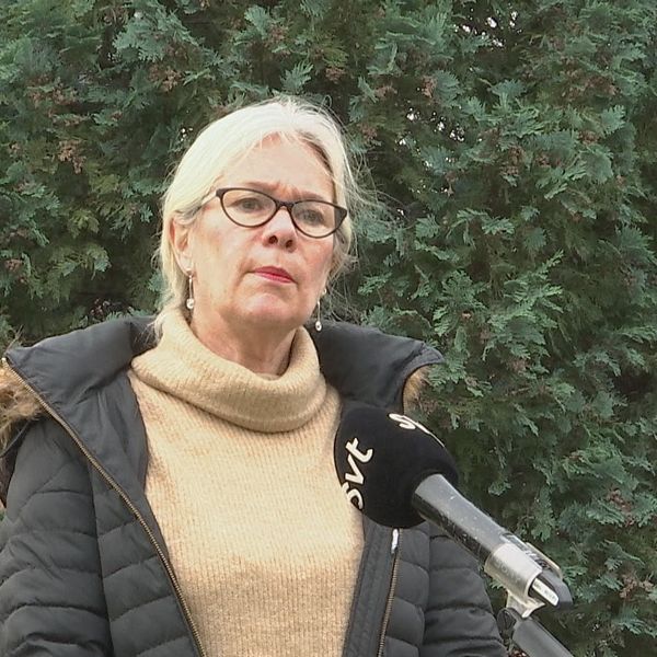 Kvinna i glasögon och tjock vinterjacka står framför en häck och pratar i en SVT-mikronfon