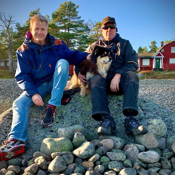 Göran Gabrielsson och Ove Olsson – två män med en hund sitter på sten i skärgården, sommarhus i bakgrund
