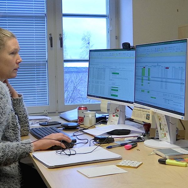 Kvinna med blont uppsatt hår sitter vid ett skrivbord med två dataskärmar.