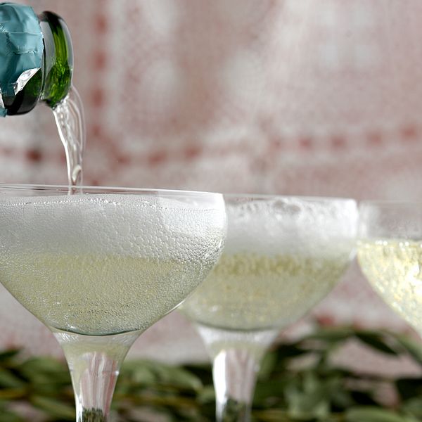 Bilden föreställer tre stycken coupeglas med mousserande vin i. I det vänstra hörnet av bilden syns en grön flaskhals med turkos folie på. Någon häller mousserande vin från flaskan och ner i det vänsta coupeglaset.