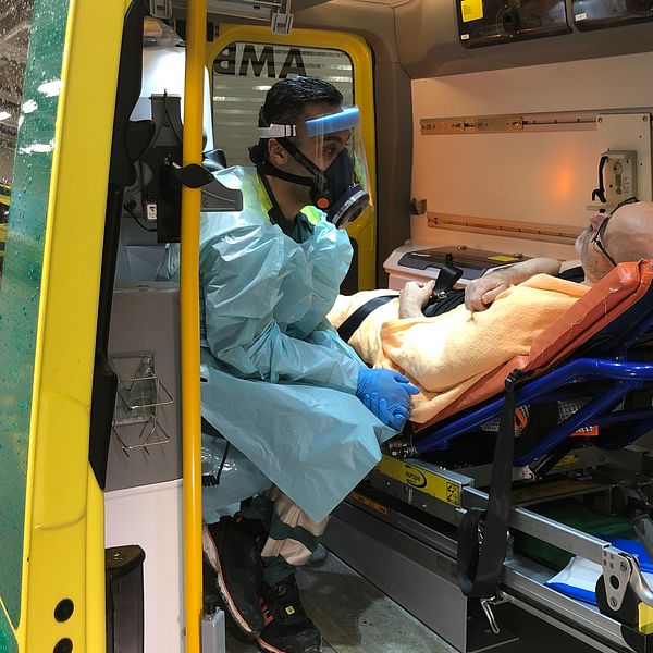En vårdanställd i skyddsutrustning sitter bredvid en patient på brits i en ambulans.