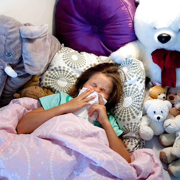 En sjuk flicka ligger nedbäddad i sängen bland en massa gossedjur och håller för näsan med en näsduk medan hon nyser.