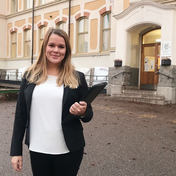 Josefin Korsbäck i svart kavaj står framför Engelska skolan i Nyfors. Hon håller en pärm i ena handen och i bakgrunden syns en basketkorg.