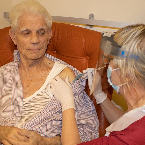 Bilden föreställer 88-årige Rune Rosander. Han sitter i en brun skinnfåtölj med händerna knäppta. Han tittar på sjuksköterskan Eva Yngvesson som injicerar covidvaccin i hans arm.