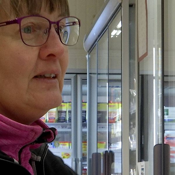Kvinna med gråsprängt uppsatt hår tittar in i kylskåp på en affär. Hon har lilafärgade bågar på glasögonen, en rosa tröja och svart jacka.