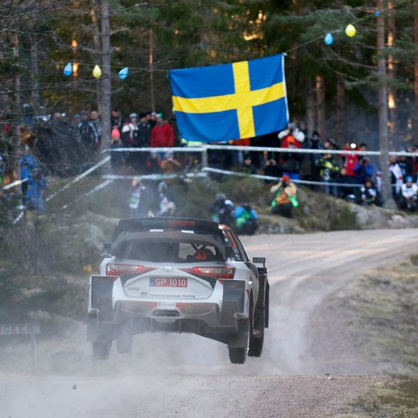 En rallybil i skogen. I bakgrunden publik samt en stor svensk flagga.