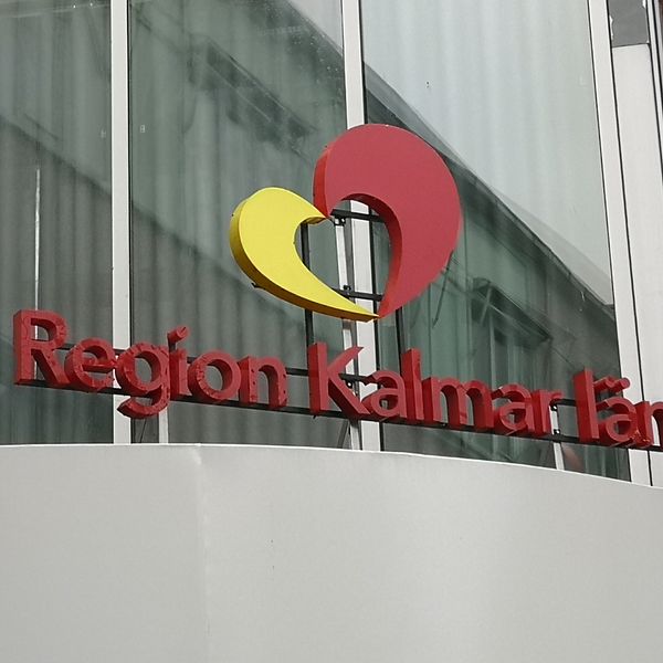 En bild på region Kalmar läns entréskylt utanför regionhuset. Det står Region Kalmar län med röda bokstäver och ovanför är deras logotyp. Ett hjärta i rött och gult.