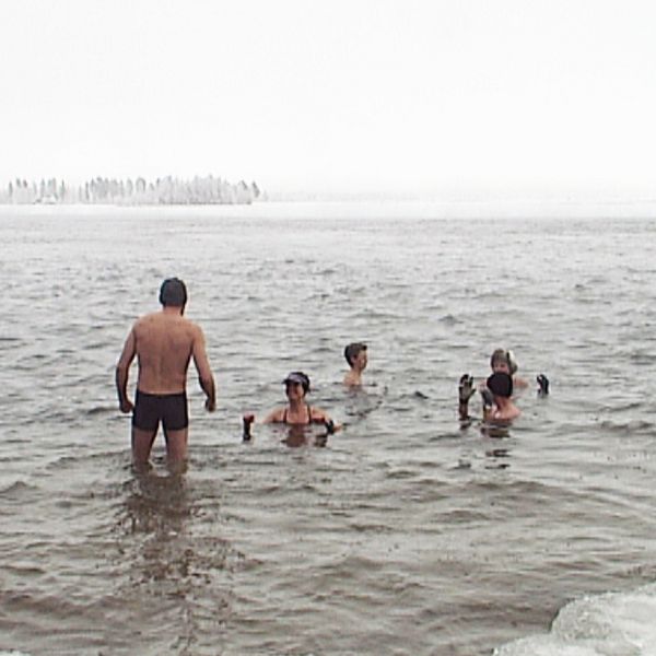 Flera personer som badar ifrån istäckt strand, snöig skog bortanför vattnet