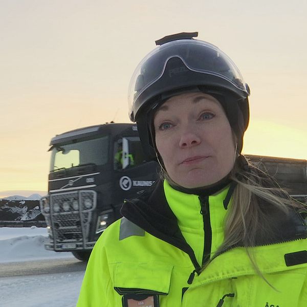 en kvinna i hjälm och varselkläder utomhus vintertid, lastbil i bakgrunden