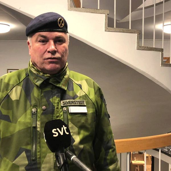 en medelålders man i militärens kamouflagekläder och basker intervjuas framför en trappa