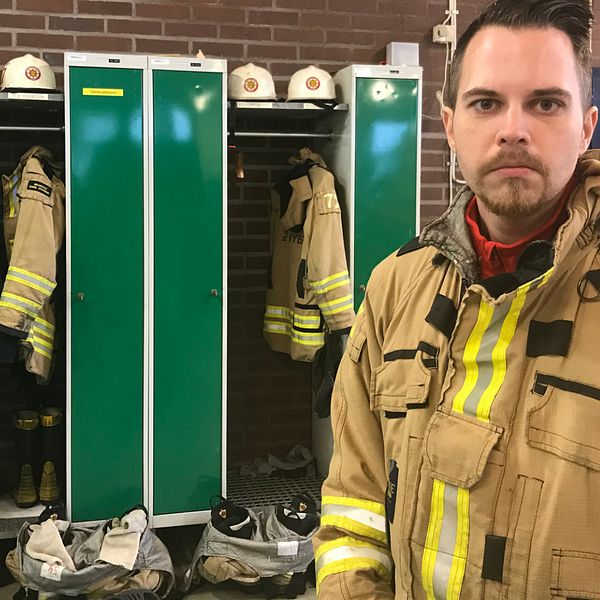 En brandman som ser ut att vara i 35-årsåldern står iklädd sin utrustning. Han står till höger bild och tittar rakt in i kameran. Han har brunt kortklippt hår, bruna ögon och skägg. Bakom honom syns gröna klädskåp och brandmansjackor som hänger på galgar. På hyllorna mellan skåpen ligger brandhjälmar.