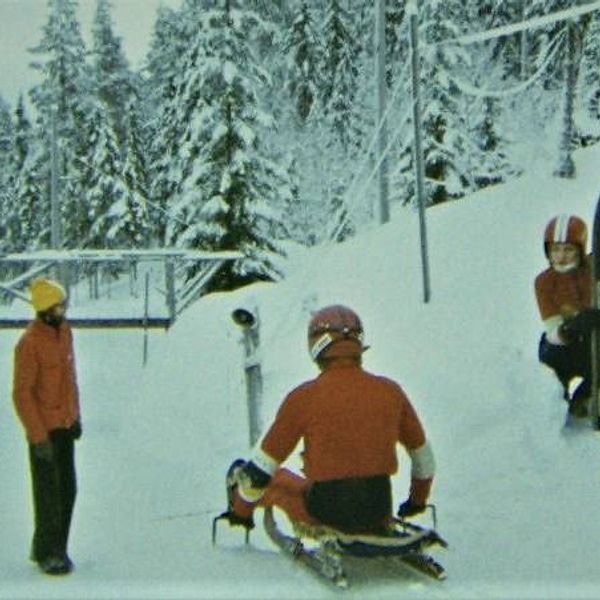 Tre personer står vid en skidbacke täckt av snö, med en skidlift i bakgrunden. En person, iklädd blå dräkt och hjälm, förbereder sig för att åka. Två andra personer, en i röd tröja och en i svart, sitter på en skida. En fjärde person, klädd i orange, står något längre bort och tittar mot gruppen. Träden i bakgrunden är täckta av snö och himlen är grå.